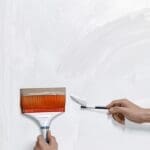Primer plano de un pincel aplicando pintura sobre una pared blanca en un entorno moderno.