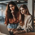 Dos emprendedoras jóvenes trabajando juntas frente a una laptop en un espacio de coworking moderno.